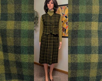 May include: A vintage green and black plaid skirt suit on a mannequin. The suit includes a knee-length skirt, a button-up vest, and a long-sleeved turtleneck sweater. The outfit is accessorized with brown shoes and earrings.
