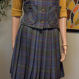 Pu&ograve; includere: Un tailleur gonna vintage a quadri su un manichino. Il completo presenta una gonna a pieghe, un gilet abbottonato con tasche e un maglione giallo senape. Il completo &egrave; impreziosito da una spilla a forma di fiore e tacchi marroni.