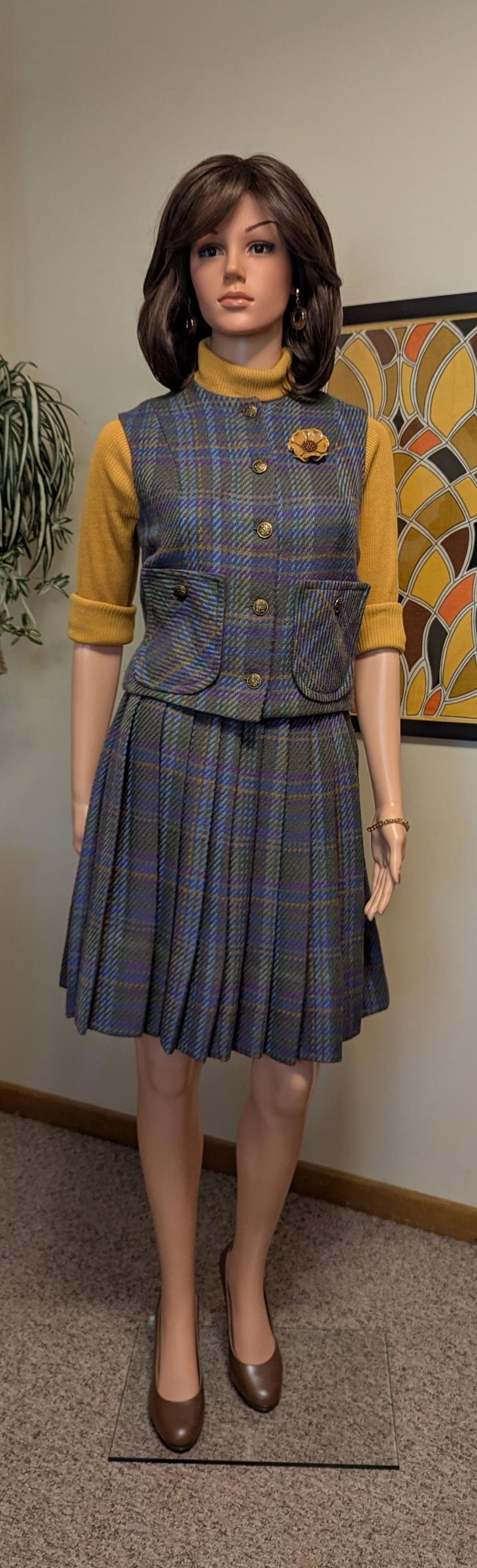 Pu&ograve; includere: Un tailleur gonna vintage a quadri su un manichino. Il completo presenta una gonna a pieghe, un gilet abbottonato con tasche e un maglione giallo senape. Il completo &egrave; impreziosito da una spilla a forma di fiore e tacchi marroni.
