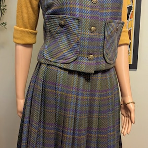 Pu&ograve; includere: Un tailleur gonna vintage a quadri con gilet e gonna a pieghe abbinati. Il gilet presenta bottoni dorati e tasche. L'outfit &egrave; un mix di tonalit&agrave; blu, verdi e viola, con un top a maniche lunghe giallo senape sottostante. Una spilla a forma di fiore dorato adorna il gilet.
