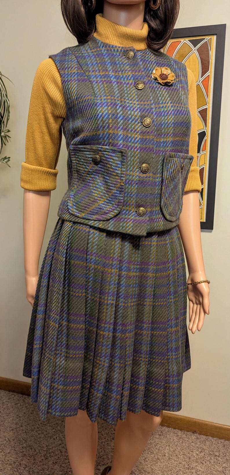 Pu&ograve; includere: Un tailleur gonna vintage a quadri con gilet e gonna a pieghe abbinati. Il gilet presenta bottoni dorati e tasche. L'outfit &egrave; un mix di tonalit&agrave; blu, verdi e viola, con un top a maniche lunghe giallo senape sottostante. Una spilla a forma di fiore dorato adorna il gilet.