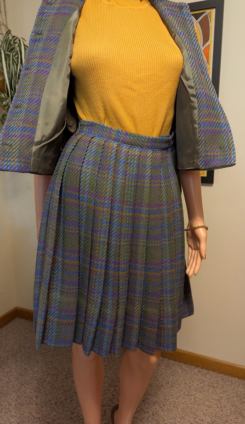 Pu&ograve; includere: Un completo gonna vintage a quadri, con gonna plissettata e giacca abbinata. La combinazione di colori comprende verde, blu, viola e giallo. L'outfit &egrave; abbinato a un top a coste giallo senape.