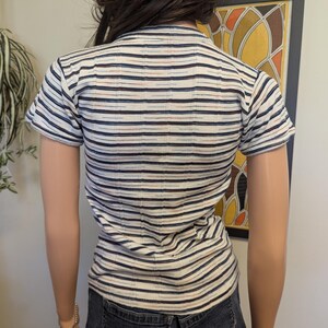 May include: A white striped short-sleeved shirt with blue, brown, and red stripes. The shirt is fitted and has a round neckline.