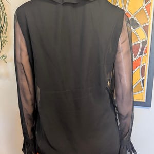 May include: Black sheer blouse with a ruffled collar and sleeves. The blouse has long sleeves with ruffled cuffs. The model is wearing an orange skirt. The background includes a framed abstract art piece.