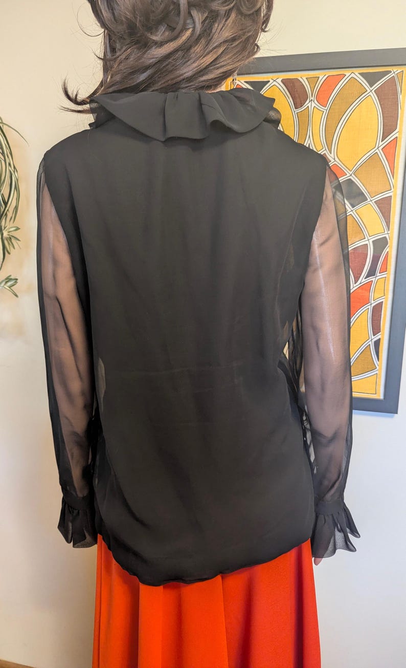 May include: Black sheer blouse with a ruffled collar and sleeves. The blouse has long sleeves with ruffled cuffs. The model is wearing an orange skirt. The background includes a framed abstract art piece.
