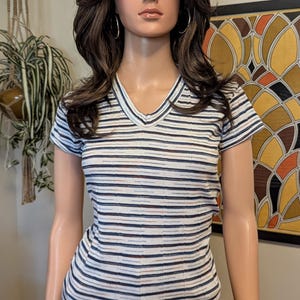 May include: A white striped v-neck t-shirt with short sleeves. The shirt has thin black, blue, and brown stripes. The shirt is being worn by a mannequin.