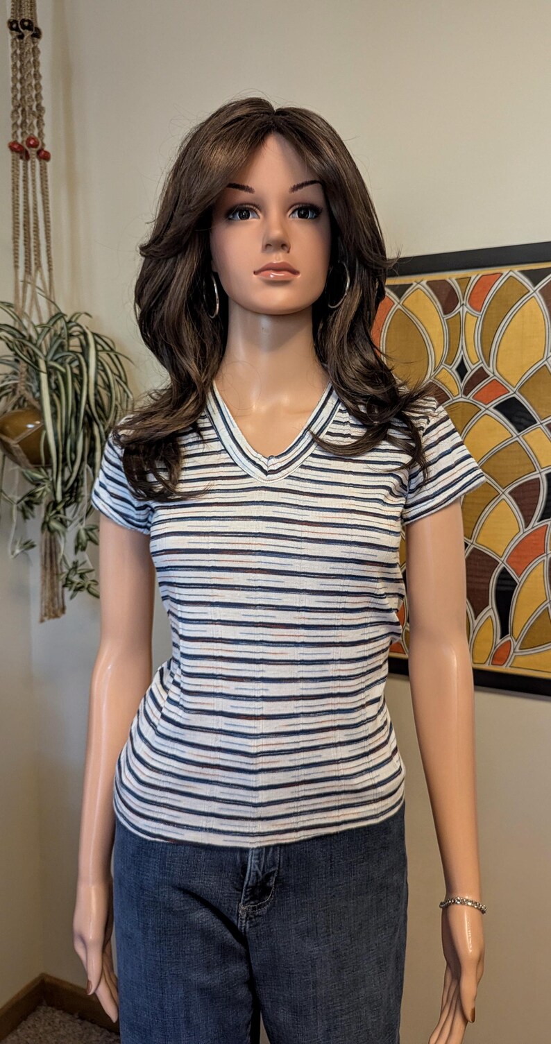 May include: A white striped v-neck t-shirt with short sleeves. The shirt has thin black, blue, and brown stripes. The shirt is being worn by a mannequin.