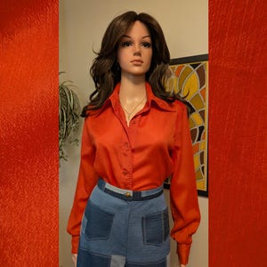 May include: A long-sleeved, button-down shirt in a vibrant orange hue, featuring a pointed collar and a smooth, slightly reflective fabric texture. The shirt is paired with a high-waisted, multi-toned blue denim skirt with visible patch pockets.