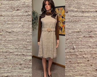 May include: A vintage-style dress on a mannequin. The dress is a light beige color with a textured pattern, a sleeveless design, and a matching belt. It is paired with a long-sleeved brown top underneath. The mannequin is wearing brown heels.