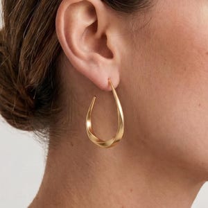 Könnte beinhalten: Ein goldener Creolen-Ohrring mit gedrehtem, tropfenförmigem Design. Der Ohrring hat eine glatte, matte Metalloberfläche und eine durchgehend geschwungene Struktur, die sich nach oben hin zum Ohrläppchen verjüngt und so einen eleganten Look erzeugt.