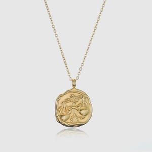 May include: Gold necklace with a zodiac pendant featuring the Libra symbol. The pendant is a round, textured disc with a raised image of the scales of justice.