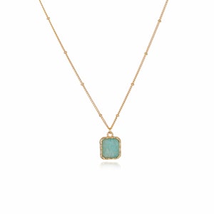 May include: A gold-colored necklace with a rectangular, light blue pendant. The pendant is framed by a textured gold border and hangs from a delicate chain. The chain features small, evenly spaced gold beads.