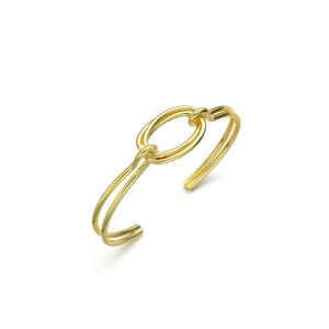 Könnte beinhalten: Ein goldfarbenes Armreif-Armband mit einem ovalen Mittelstück. Das Armband hat ein Doppelband-Design, das sich um das Handgelenk krümmt. Das Oval ist mit einem knotenartigen Detail mit dem Band verbunden. Schmuck.