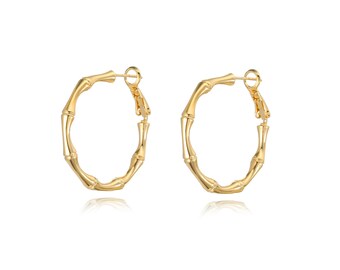May include: A pair of gold-colored hoop earrings. The earrings have a bamboo-like design, with segmented sections creating a textured appearance. The hoops are circular and have a clasp closure. The earrings are displayed against a white background.