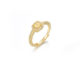 May include: A gold ring featuring a square yellow gemstone set within a halo of smaller clear stones.