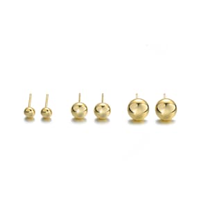 May include: Six pairs of gold-colored ball stud earrings in varying sizes are displayed against a white background. The earrings are round and shiny, reflecting light. The sizes range from small to large, showcasing a simple, classic design.