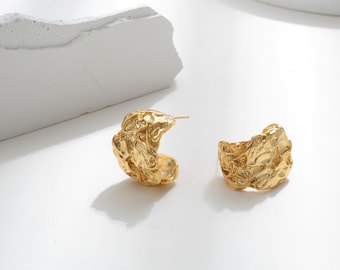 May include: A pair of gold-toned hoop earrings with a textured, irregular surface. The earrings are made of metal and have a chunky, modern design.