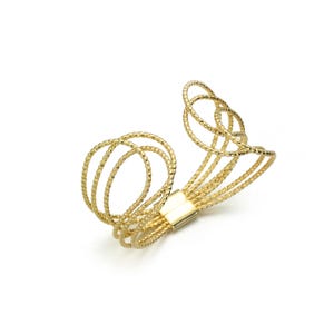 May include: A gold-colored arm cuff bracelet. The bracelet features multiple textured, looped strands that connect at the center with a rectangular clasp. The design is open and airy, with a focus on the interwoven strands.