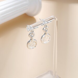 May include: A pair of silver dangle earrings with a textured circular design. The earrings are displayed on a clear acrylic stand.