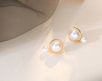 Könnte beinhalten: Ein Paar goldfarbene Ohrringe mit einem Perlenanhänger und einem kleineren Perlenakzent. Die Ohrringe liegen auf einer weißen Oberfläche.