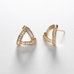 May include: A pair of gold-colored triangle stud earrings. Each earring features a white, curved inlay and is encrusted with small, clear rectangular stones. The earrings have a post-and-clutch closure.