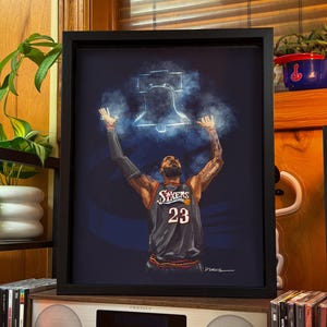 May include: A framed art print featuring a basketball player in a black Sixers jersey with the number 23, arms raised toward a glowing blue Liberty Bell illustration against a dark background. The artwork includes a signature in the bottom right corner.