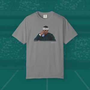 May include: A gray t-shirt featuring a graphic of a person drinking from a can. The person is wearing a headband and a dark green sweater. The image is a drawing.