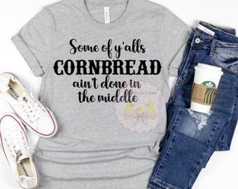 May include: A gray t-shirt with the text "Some of y'alls Cornbread ain't done in the middle" printed in black. The shirt is laid flat on a white surface.