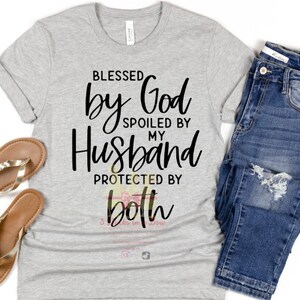 May include: A gray t-shirt with the text "Blessed by God Spoiled by My Husband Protected by Both" in black lettering. The shirt is shown on a white background with a pair of brown sandals and blue jeans.