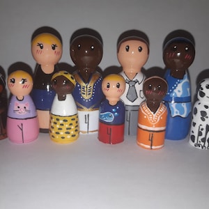 Puede incluir: Un conjunto de nueve muñecos de madera pintados a mano, incluyendo un perro y un gato. Los muñecos están vestidos con diferentes atuendos coloridos, incluyendo un vestido azul, un vestido amarillo con lunares negros, una camisa azul y dorada, una camisa roja con un diseño de pez, una camisa gris con corbata, una camisa azul y blanca, una camisa naranja con el número 4 y una camisa azul y blanca con un diseño floral.