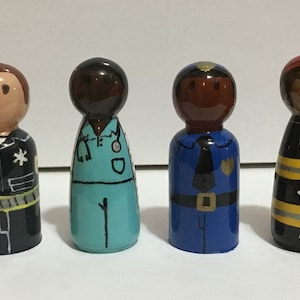 Peut inclure: Quatre poupées en bois peintes pour représenter différentes professions : un policier, un médecin, un policier et un pompier. Les poupées sont toutes debout sur une surface blanche.