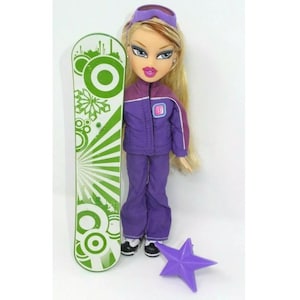 Puede incluir: Una muñeca rubia con un traje de esquí morado y gafas, sosteniendo una tabla de snowboard verde y blanca con un accesorio en forma de estrella.