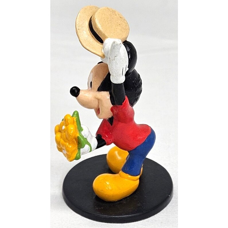 Puede incluir: Una colorida figura de Mickey Mouse sosteniendo un ramo de flores amarillas y un sombrero de paja. Mickey lleva una camisa roja, pantalones azules y zapatos amarillos. La figura est&aacute; sobre una base circular negra.