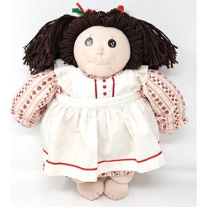 Op de afbeelding: Een handgemaakte stoffen pop met bruin garen haar, een witte jurk met rode bies en een roze en wit gestreept shirt met aardbeien. De pop heeft één zwart oog en één grijs oog.