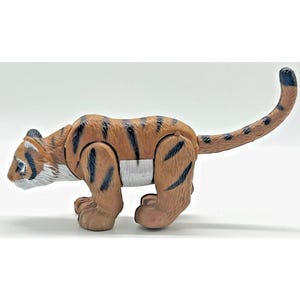 Puede incluir: Una figura de tigre de plástico en postura de acecho, con cuerpo color canela, rayas verticales negras, barbilla y pecho blancos, y una cola larga y curvada. El juguete tiene puntos de articulación visibles en los hombros y las caderas, mostrando un diseño segmentado.