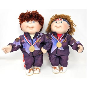 May include: Two Cabbage Patch Kids dolls dressed in purple and blue Olympic tracksuits with gold medals. The dolls have red and brown hair and are wearing white sneakers.