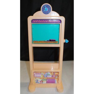 May include: A pink plastic toy schoolhouse with a chalkboard, a clock, and a bookshelf. The chalkboard has a green surface and the alphabet is printed above it. The clock has a blue face and black numbers. The bookshelf has a picture of books, pencils, and other school supplies.