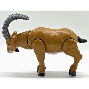 May include: A brown plastic ibex figurine standing in profile. The animal features a textured coat, a small black beard, and large, curved, ridged grey horns. It has black hooves and a short black tail, with visible articulation joints at the shoulders.