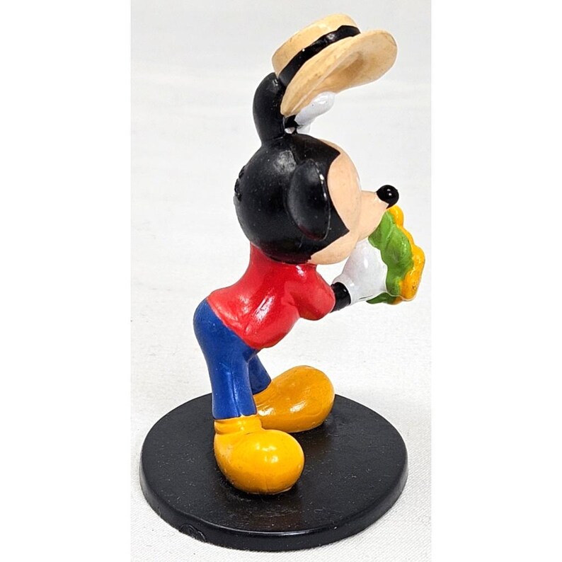 Puede incluir: Una figura de Mickey Mouse en una pose cl&aacute;sica, con camisa roja, pantalones azules y zapatos amarillos. El personaje de orejas negras sostiene un objeto amarillo y verde y lleva un sombrero de paja. La figura est&aacute; sobre una base circular negra.