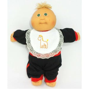 Op de afbeelding: Een Cabbage Patch Kid pop met blond haar, groene ogen en een roze blos. De pop draagt een zwart en rood outfit met een wit slabbetje met een gele geborduurde giraffe.