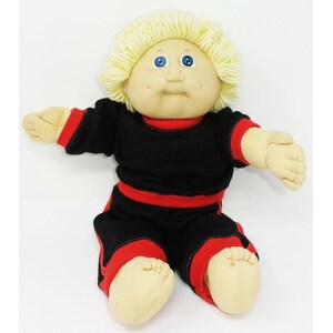 Könnte beinhalten: Eine Cabbage Patch Kid Puppe mit blonden Haaren und blauen Augen, die einen schwarzen Trainingsanzug mit roten Streifen trägt.