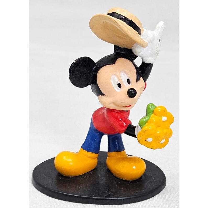 Puede incluir: Una figura de Mickey Mouse, con el ic&oacute;nico personaje con una camisa roja, pantalones azules y zapatos amarillos. Mickey sostiene una fruta amarilla y un sombrero beige. La figura est&aacute; sobre una base negra.