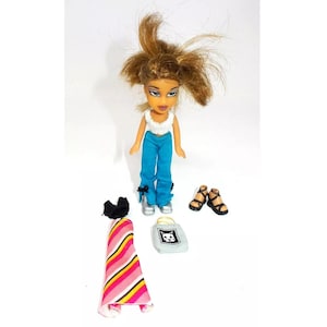 May include: A doll with brown hair and blue jeans, wearing a white shirt and holding a small purse. The doll is wearing sandals and has a pink and yellow striped dress next to it.