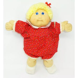 Può includere: Una bambola Cabbage Patch Kid con capelli biondi, occhi azzurri e un vestito rosso a fiori. La bambola indossa calzini bianchi e ha un fiocco rosso nei capelli.