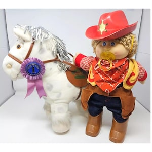 Pode incluir: Uma boneca Cabbage Patch Kid vestida de caubói montando um cavalo de pelúcia branco. A boneca está usando um chapéu de caubói vermelho com uma estrela de xerife, uma camisa xadrez vermelha e branca, um lenço vermelho, um colete amarelo, jeans azul, botas de caubói marrons e uma chupeta amarela. O cavalo tem uma fita roxa com uma roseta azul de '1º lugar'.