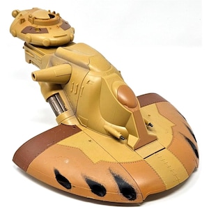 May include: A tan and brown toy vehicle with a rounded front and a raised top section. The vehicle has a sleek design with detailed features, including a central cockpit and side-mounted cannons. The underside has black paint marks.