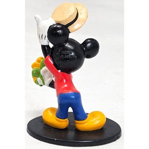 Puede incluir: Figura de Mickey Mouse, vista desde atr&aacute;s, sosteniendo un ramo de flores y un sombrero de paja. Mickey lleva una camisa roja, pantalones azules y zapatos amarillos. La figura est&aacute; sobre una base negra.