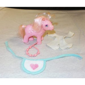 May include: A pink pony with a light blue bib, a white dress, and a pink and white beaded necklace. The pony has a pink heart on its bib.