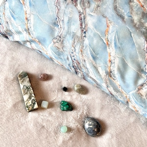 May include: A collection of various stones and crystals, including a long, green and white stone, a small green stone, a small white stone, a small black stone, a small gray stone, a small pink stone, a small green stone, and a large gray stone with gold flecks.
