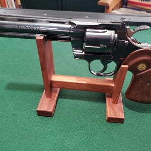 Può includere: Un revolver in metallo nero con una canna lunga contrassegnata Python 357, dotato di un'impugnatura in legno marrone testurizzato con un emblema circolare dorato, poggiato su un supporto espositivo in legno a forma di H con due montanti.